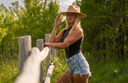 A woman in a cowboy hat, black sleeveless top, and denim shorts leans against a wooden fence in a grassy outdoor setting with trees in the background.