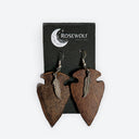 A pair of rustic dark brown wooden arrowhead dangle earrings with small silver feather charms, displayed on a black Rosewolf Designs brand card against a light gray background.
